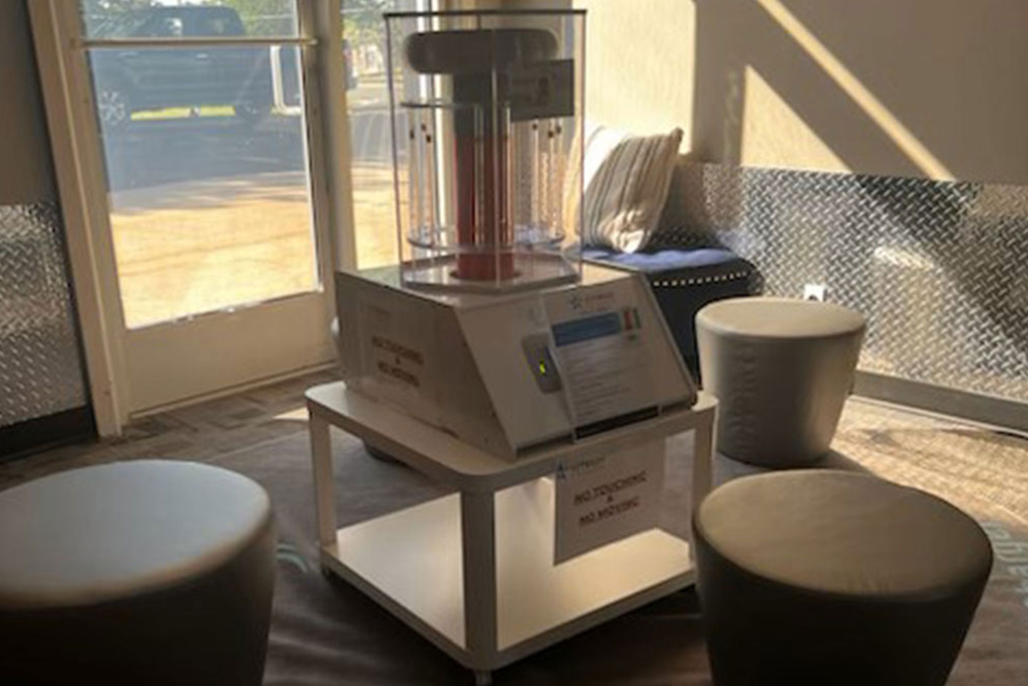 Biocharger A medium-sized disinfecting machine, encased in a clear box, sits on a white table in a well-lit room with a window, seating, and metallic wall detailing.