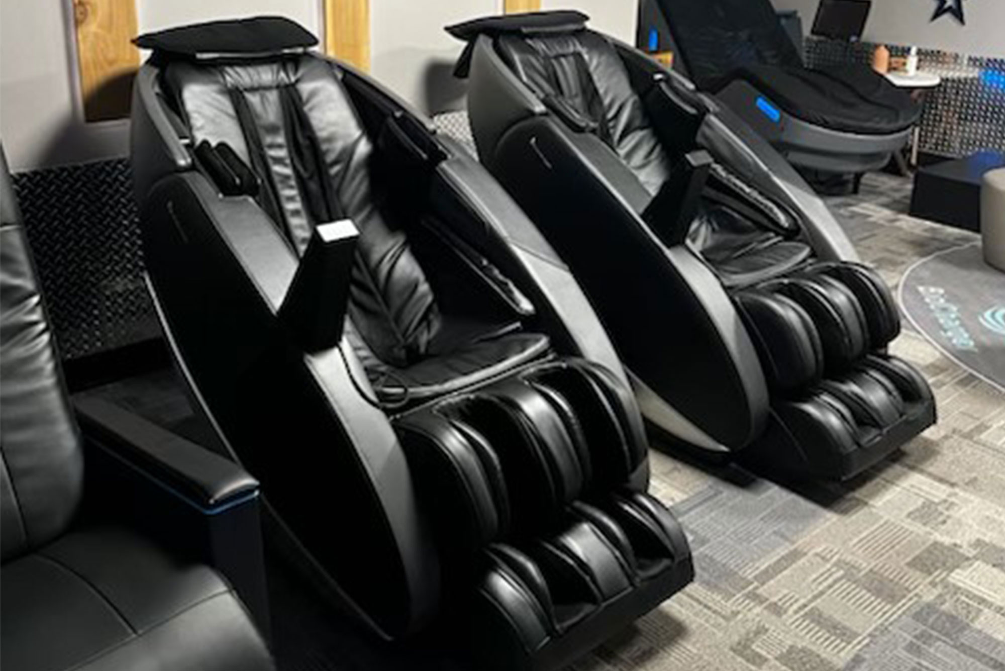 Humantouch-Massage-Chair Two black massage chairs are positioned side by side in a modern room with carpeted flooring and a sleek design. The chairs feature multiple massage rollers and footrests.