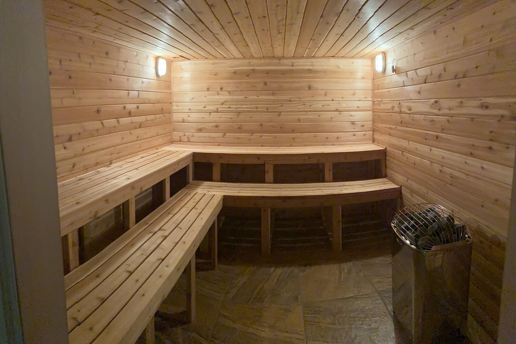 Sauna An empty, wooden sauna room with benches along the walls, lit by two wall-mounted lights, and a metal heater in the corner.