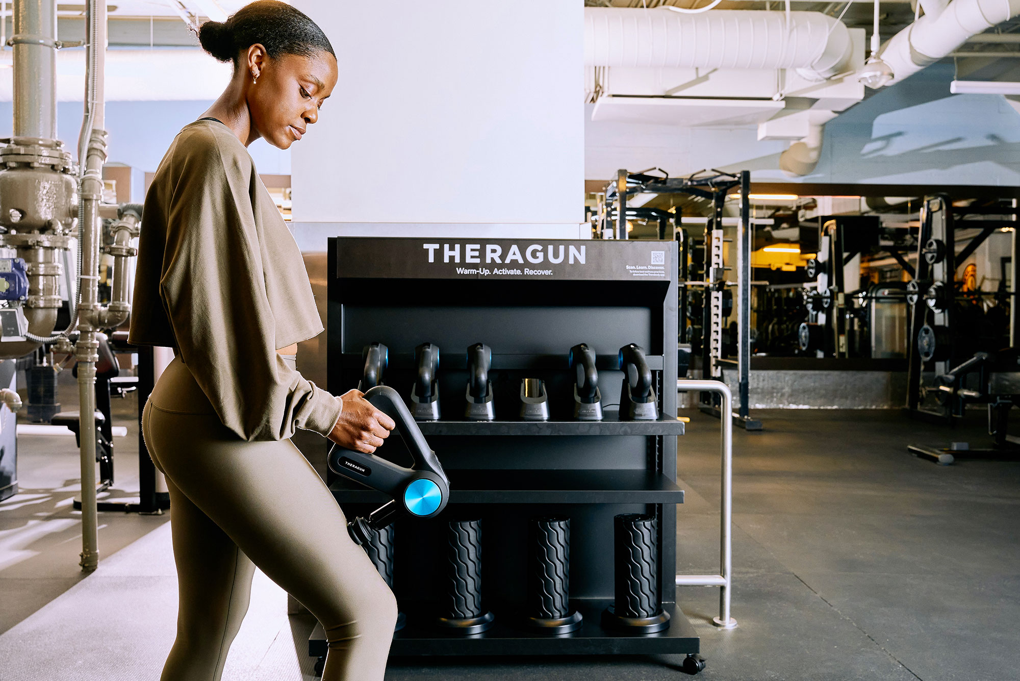Theragun A woman in athletic wear uses a Theragun in a gym, standing near a rack of other Theragun devices against a backdrop of gym equipment and fitness machines.