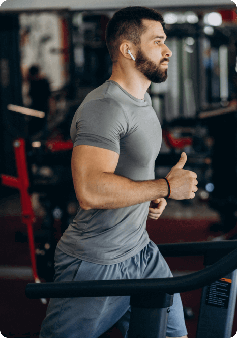 fitness-training-program Man with a beard running on a treadmill in a gym, wearing a gray shirt and earphones.